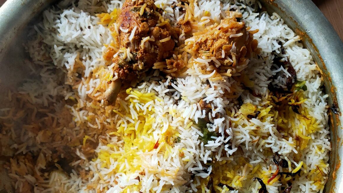 A pot of chicken dum biryani just opened, with saffron-coloured rice, fried onions and chicken pieces on top and a bowl of raita beside it